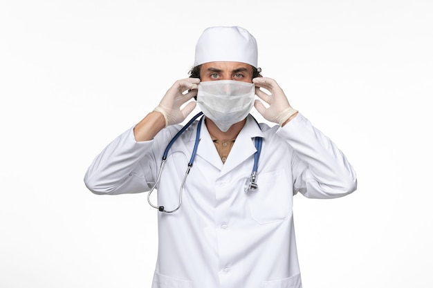 Vista frontal médico masculino en traje médico con máscara estéril como protección contra el covid- en la pared blanca enfermedad del virus covid