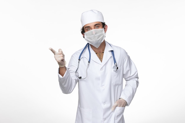 Vista frontal médico masculino en traje médico con máscara estéril como protección contra el covid- posando en la pared blanca enfermedad del virus enfermedad pandémica del covid