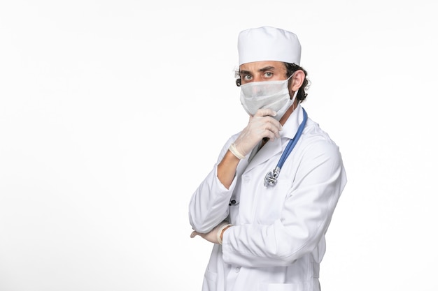 Vista frontal médico masculino en traje médico con máscara estéril como protección contra covid posando y pensando en la salud de la pandemia de coronavirus de salpicaduras de pared blanca