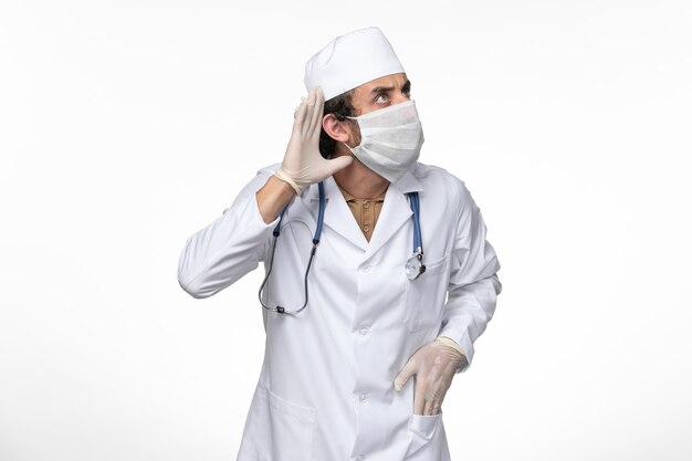 Vista frontal médico masculino en traje médico con máscara estéril como protección contra el covid- tratando de escuchar en la pared blanca enfermedad del virus enfermedad pandémica del covid