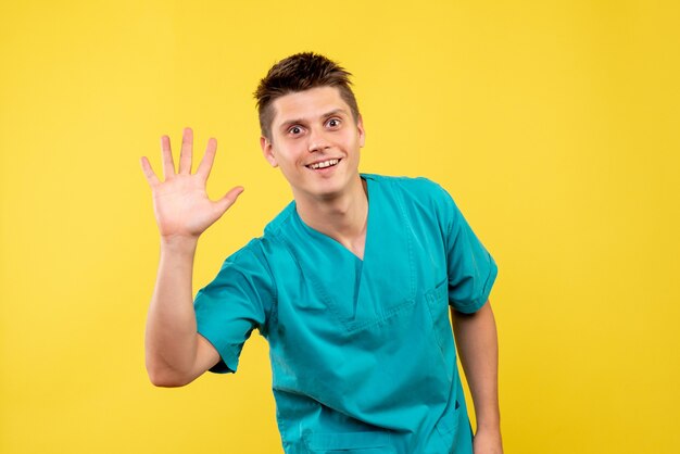 Vista frontal del médico masculino en traje médico saludo en pared amarilla