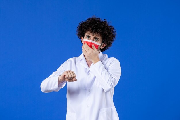 Vista frontal médico masculino en traje médico y tiempo de control de máscara sobre fondo azul vacuna hospital virus covid- medicina pandémica droga