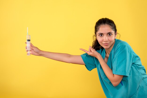 Vista frontal médico de la mujer bonita en uniforme apuntando a la jeringa de pie sobre fondo amarillo