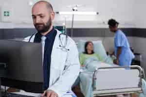 Foto gratuita vista frontal del médico practicante que escribe la experiencia médica en la computadora mientras que en segundo plano el asistente negro discute el tratamiento de la salud. paciente hospitalizado con trastorno respiratorio