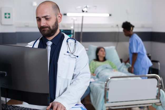 Vista frontal del médico practicante que escribe la experiencia médica en la computadora mientras que en segundo plano el asistente negro discute el tratamiento de la salud. Paciente hospitalizado con trastorno respiratorio