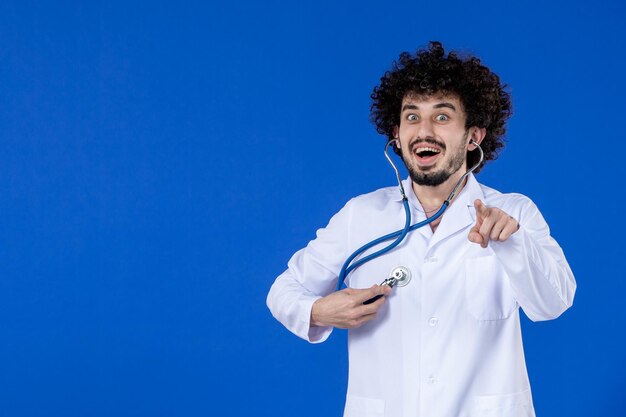 Vista frontal del médico de sexo masculino en traje médico comprobando su cuerpo con estetoscopio sobre superficie azul