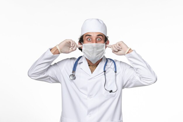 Vista frontal médico de sexo masculino en traje médico con máscara como protección contra el covid: cierre de las orejas en el escritorio blanco medicina pandémica del virus del coronavirus
