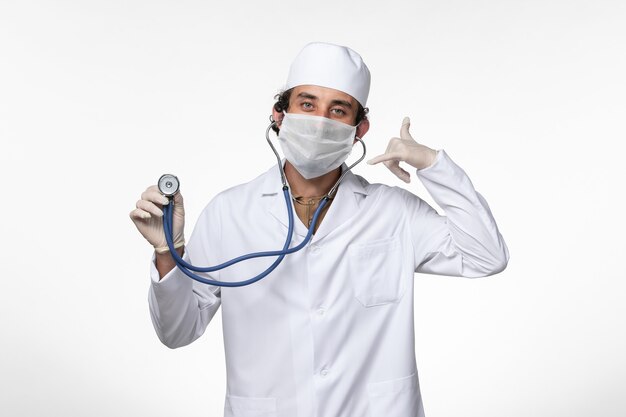 Vista frontal médico de sexo masculino en traje médico y con una máscara como protección contra el covid - usando estetoscopio en la pared blanca enfermedad covid - pandemia