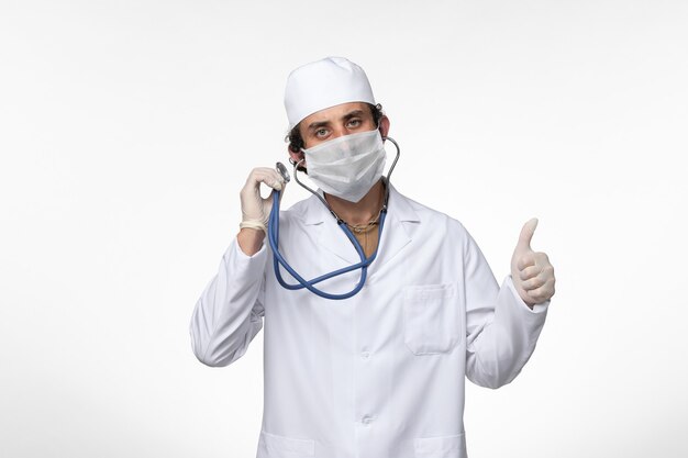 Vista frontal médico de sexo masculino en traje médico y con una máscara como protección contra el covid - usando estetoscopio en la pared blanca enfermedad covid - pandemia