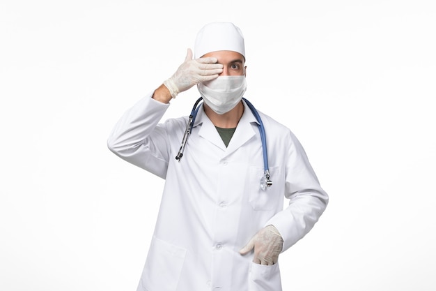 Vista frontal médico de sexo masculino en traje médico y con una máscara contra el covid con estetoscopio azul en la pared blanca clara covid- enfermedad enfermedad pandémica
