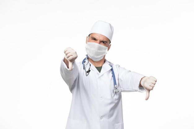 Vista frontal médico de sexo masculino en traje médico y con una máscara contra covid con estetoscopio en la pared blanca covid- enfermedad enfermedad pandémica virus