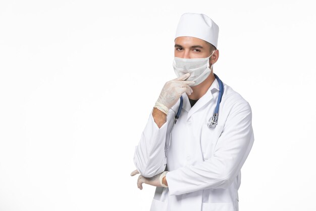 Vista frontal médico de sexo masculino en traje médico y con máscara debido al covid-pensamiento en la pared blanca covid-virus enfermedad pandémica de medicina