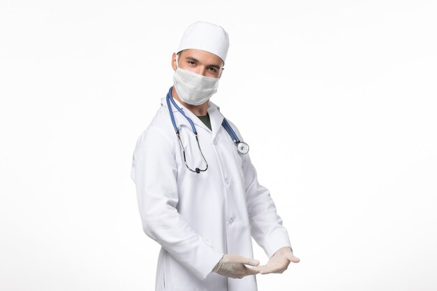 Vista frontal médico de sexo masculino en traje médico y con máscara debido a covid- posando en la pared blanca covid- virus pandémico virus