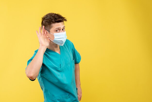 Vista frontal del médico de sexo masculino en traje médico con máscara estéril escuchando en la pared amarilla