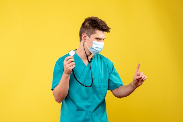 Vista frontal del médico de sexo masculino en traje médico y máscara con estetoscopio en pared amarilla