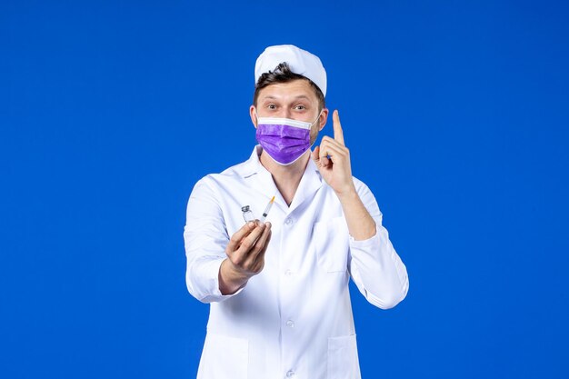 Vista frontal del médico de sexo masculino en traje médico y máscara con inyección y vacuna en azul