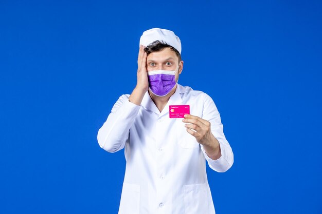 Vista frontal del médico de sexo masculino en traje médico y máscara con tarjeta de crédito en azul