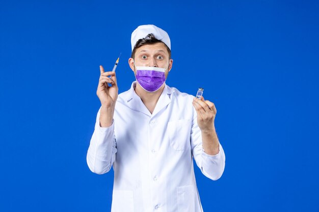 Vista frontal del médico de sexo masculino en traje médico y máscara con vacuna e inyección en azul