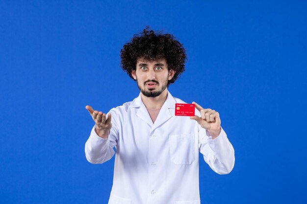 Vista frontal del médico de sexo masculino en traje médico con tarjeta de crédito roja sobre superficie azul