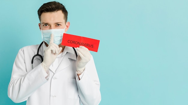 Vista frontal del médico sosteniendo papel con coronavirus y haciendo la señal silenciosa