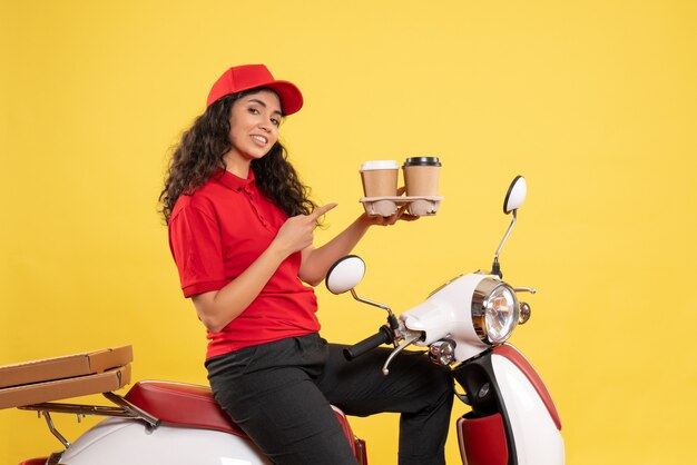 Vista frontal de mensajería femenina en bicicleta sosteniendo tazas de café sobre fondo amarillo servicio de trabajador uniforme trabajo mujer entrega trabajo