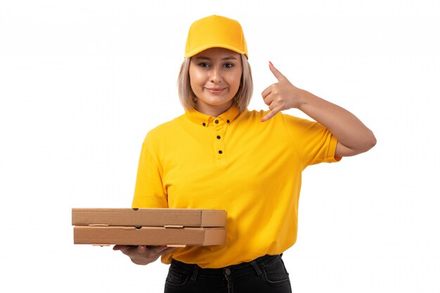Una vista frontal de mensajería femenina en camisa amarilla gorra amarilla y jeans negros con cajas de pizza mostrando señal de llamada telefónica sonriendo en blanco
