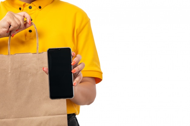 Una vista frontal de mensajería femenina en camisa amarilla gorra amarilla y jeans negros con paquete con comida y teléfono inteligente en blanco