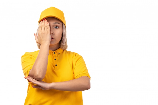 Una vista frontal de mensajería femenina en camisa amarilla gorra amarilla y jeans negros posando cubriendo su ojo en blanco