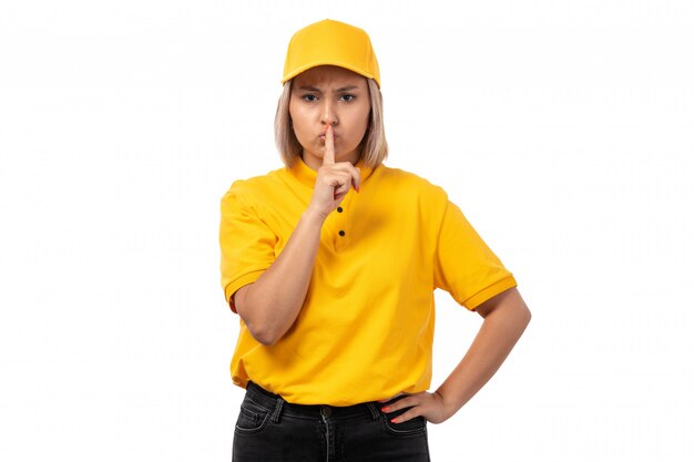 Una vista frontal de mensajería femenina en camisa amarilla gorra amarilla y jeans negros posando mostrando señal de silencio en blanco