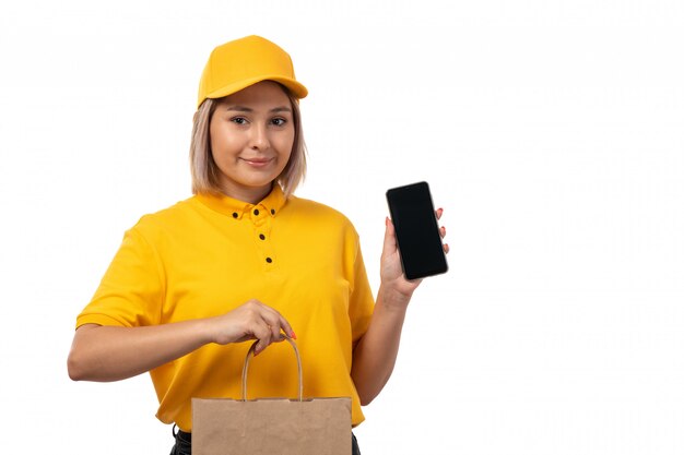 Una vista frontal de mensajería femenina en camisa amarilla gorra amarilla y jeans negros con smartphone y paquete con comida sonriendo en blanco