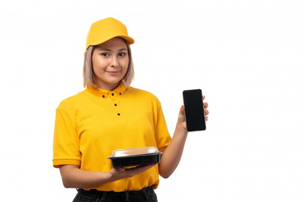 Una vista frontal de mensajería femenina en camisa amarilla gorra amarilla y jeans negros con smartphone y tazón con comida sonriendo en blanco