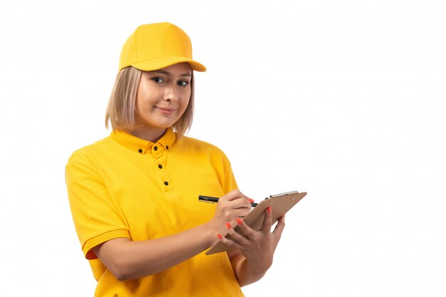 Una vista frontal de mensajería femenina en camisa amarilla gorra amarilla jeans negros sonriendo escribiendo órdenes en blanco