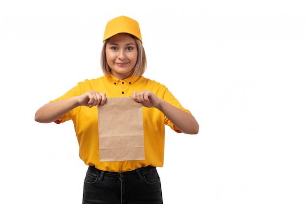 Una vista frontal de mensajería femenina en camisa amarilla gorra amarilla y jeans negros sonriendo y sosteniendo paquete de alimentos en blanco