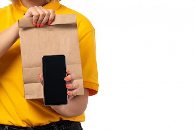 Una vista frontal de mensajería femenina en camisa amarilla gorra amarilla jeans negros sonriendo sosteniendo smartphone y paquetes con comida en blanco