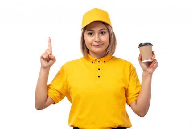 Una vista frontal de mensajería femenina en camisa amarilla gorra amarilla y jeans negros sonriendo sosteniendo la taza de café en blanco