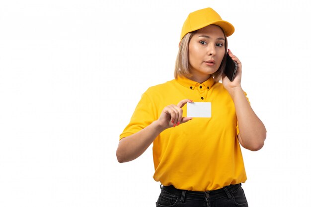 Foto gratuita una vista frontal de mensajería femenina en camisa amarilla gorra amarilla y jeans negros con tarjeta blanca hablando por teléfono en blanco