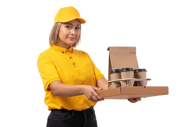 Una vista frontal de mensajería femenina en camisa amarilla gorra amarilla y jeans negros con tazas de café y caja de pizza en blanco