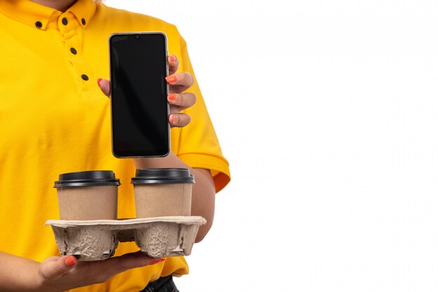 Una vista frontal de mensajería femenina en camisa amarilla gorra amarilla y jeans negros con tazas de café y teléfono inteligente en blanco