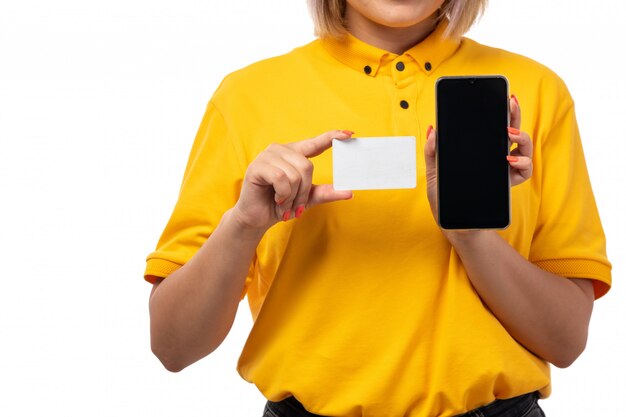 Una vista frontal de mensajería femenina en camisa amarilla gorra amarilla y jeans negros con teléfono inteligente y tarjeta blanca sobre blanco