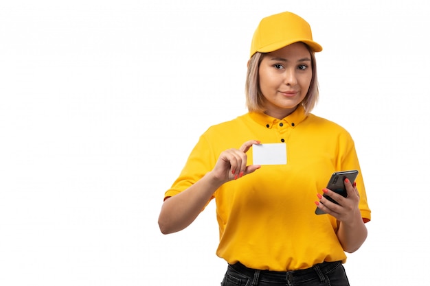 Una vista frontal de mensajería femenina en camisa amarilla gorra amarilla y jeans negros con teléfono inteligente y tarjeta blanca sonriendo en blanco