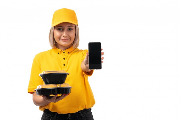 Una vista frontal de mensajería femenina en camisa amarilla y gorra amarilla sosteniendo cuencos con comida smartphone sonriendo en blanco