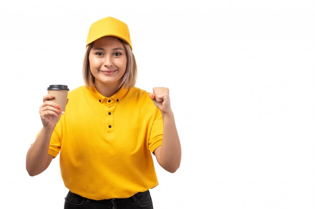 Una vista frontal de mensajería femenina en camisa amarilla gorra amarilla con taza de café sonriendo felizmente en blanco