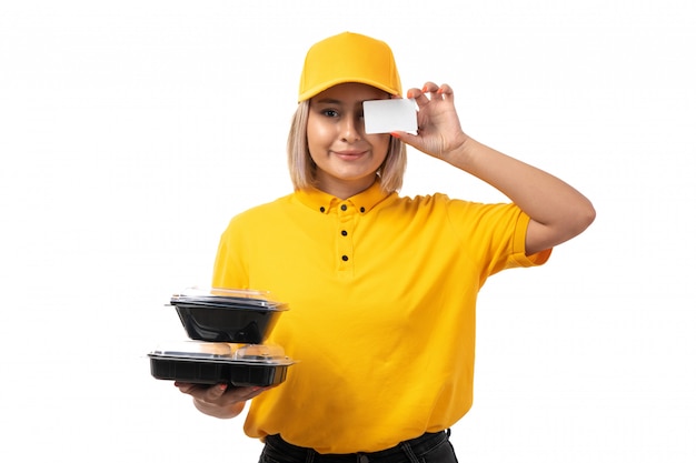 Una vista frontal de mensajería femenina en camisa amarilla tapa amarilla sosteniendo cuencos con comida y tarjeta blanca sonriendo en blanco