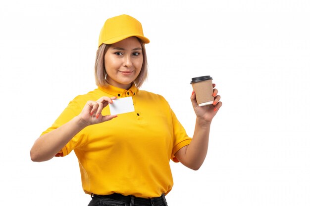 Una vista frontal de mensajería femenina en camisa amarilla yellwo gorra y jeans negros sonriendo sosteniendo una taza de café y una tarjeta blanca en uniforme blanco