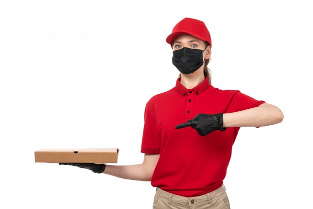 Una vista frontal de mensajería femenina en camisa roja gorra roja guantes negros y máscara negra con caja de pizza señalando en blanco