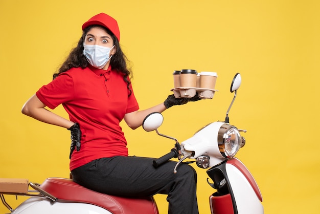 Vista frontal de mensajería femenina en máscara en bicicleta con tazas de café sobre fondo amarillo servicio de trabajador uniforme pandemia mujer de trabajo covid-