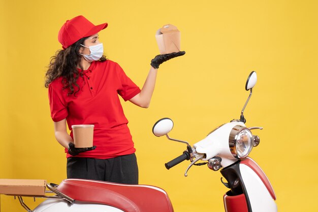 Vista frontal de mensajería femenina en máscara con entrega de postre y comida sobre fondo amarillo covid- trabajo uniforme de trabajo servicio de entrega