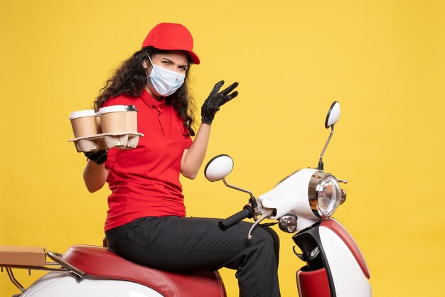 Vista frontal de mensajería femenina en máscara con tazas de café sobre fondo amarillo pandemia trabajador entrega uniforme covid- trabajo