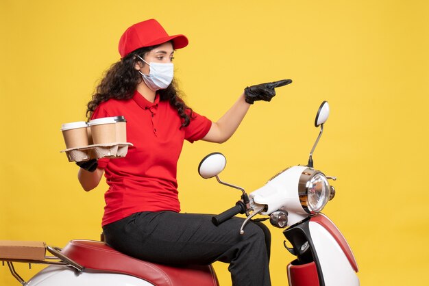 Vista frontal de mensajería femenina en máscara con tazas de café sobre fondo amarillo uniforme de trabajador de pandemia de servicio covid- entrega de trabajo