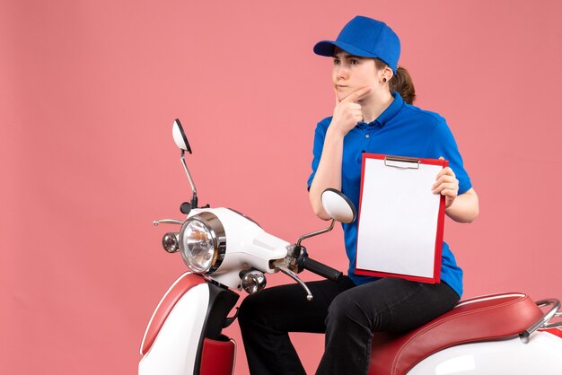 Vista frontal de mensajería femenina sentada en bicicleta con nota de archivo en color rosa uniforme de servicio de entrega de alimentos trabajador de trabajo